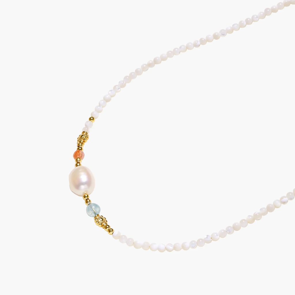 Delicate white beaded necklace with baroque pearl center, gold accents, and coral and aqua gemstones on cream background.