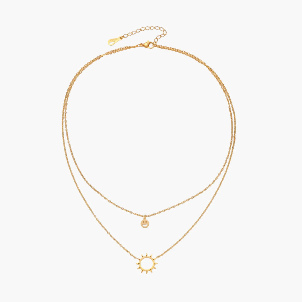 Gold layered necklace set with sunburst and smiley face pendants on delicate chains against white background.