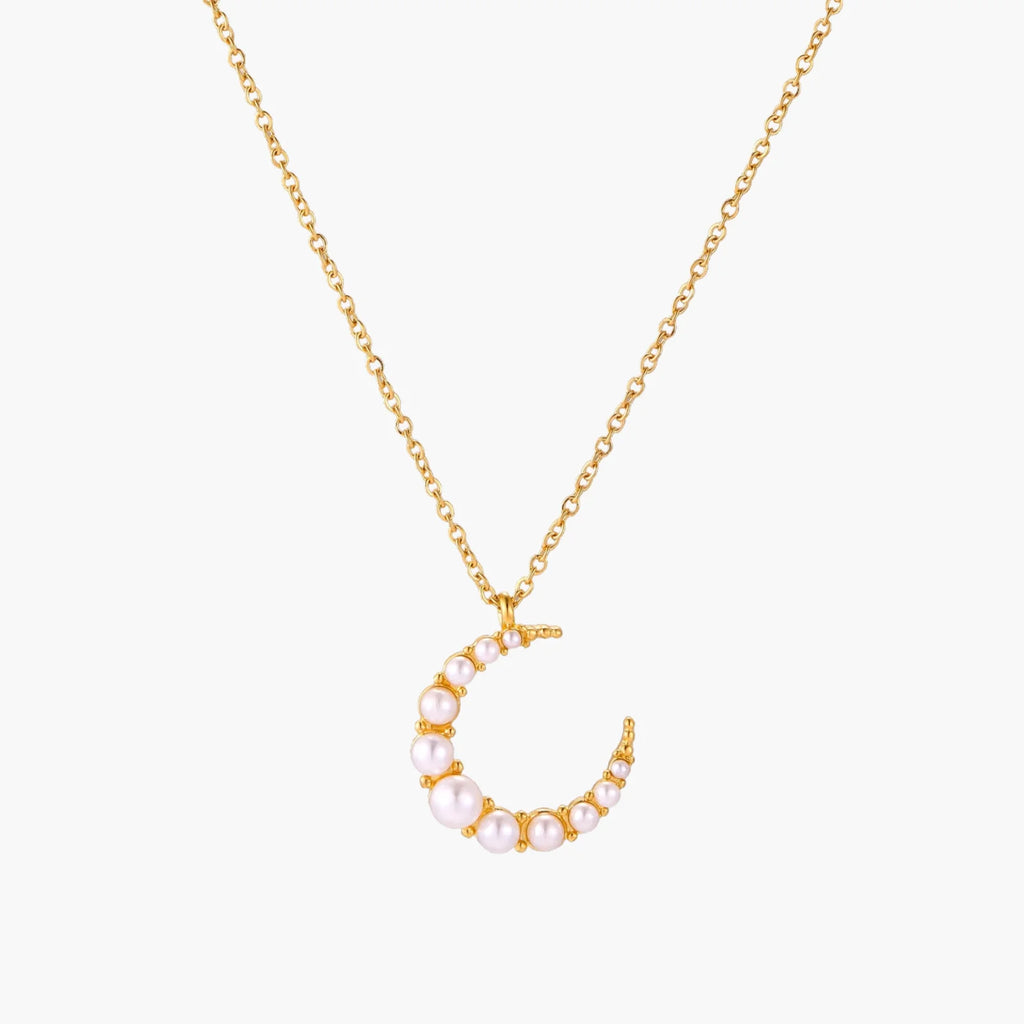 Gold crescent moon pendant with graduated white pearls on delicate chain against cream background