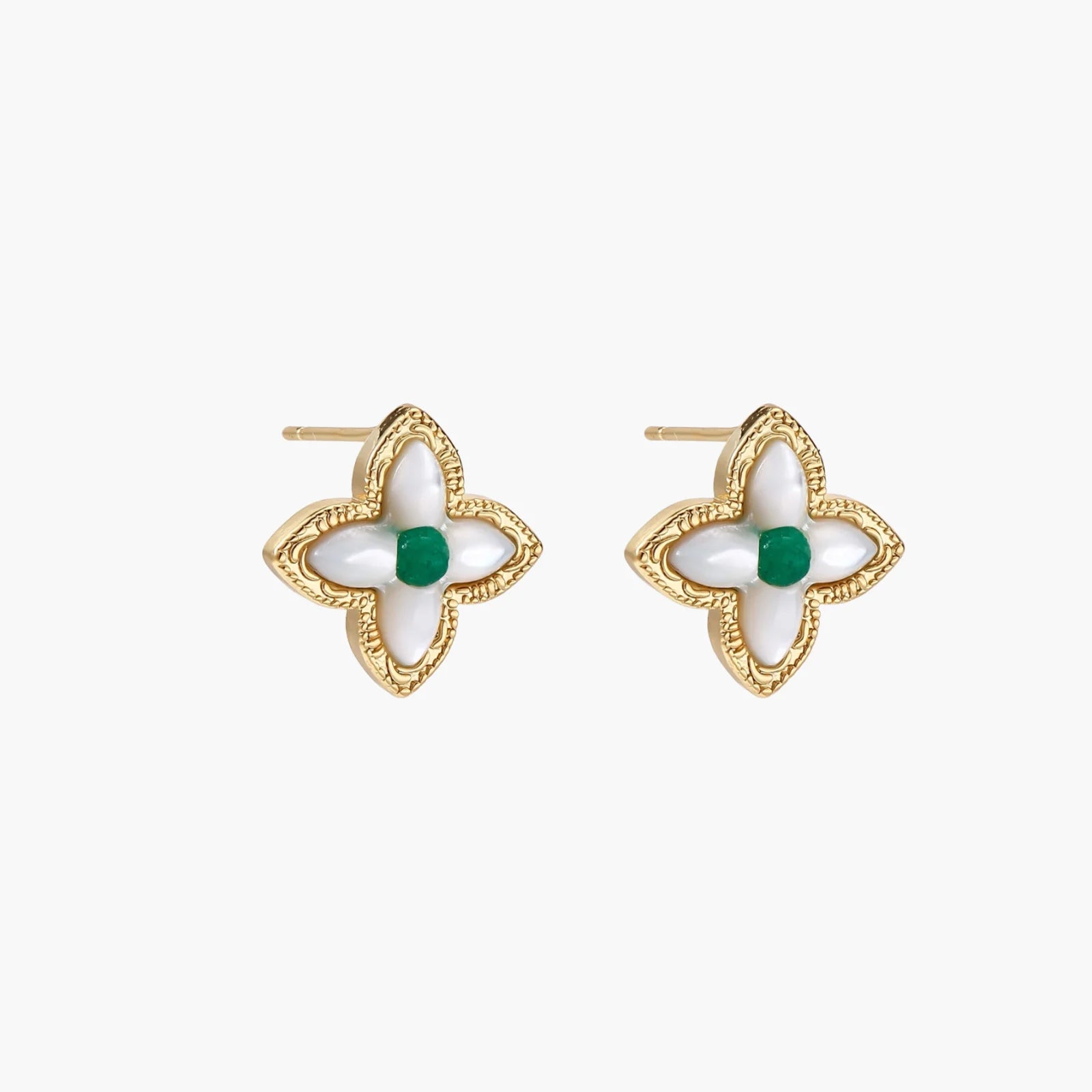 Gold quatrefoil stud earrings with white mother-of-pearl petals and emerald green gemstone center on beige background.