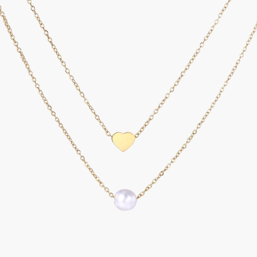 Two delicate gold-plated chains with heart charm and white pearl bead on cream background, minimal layered design.