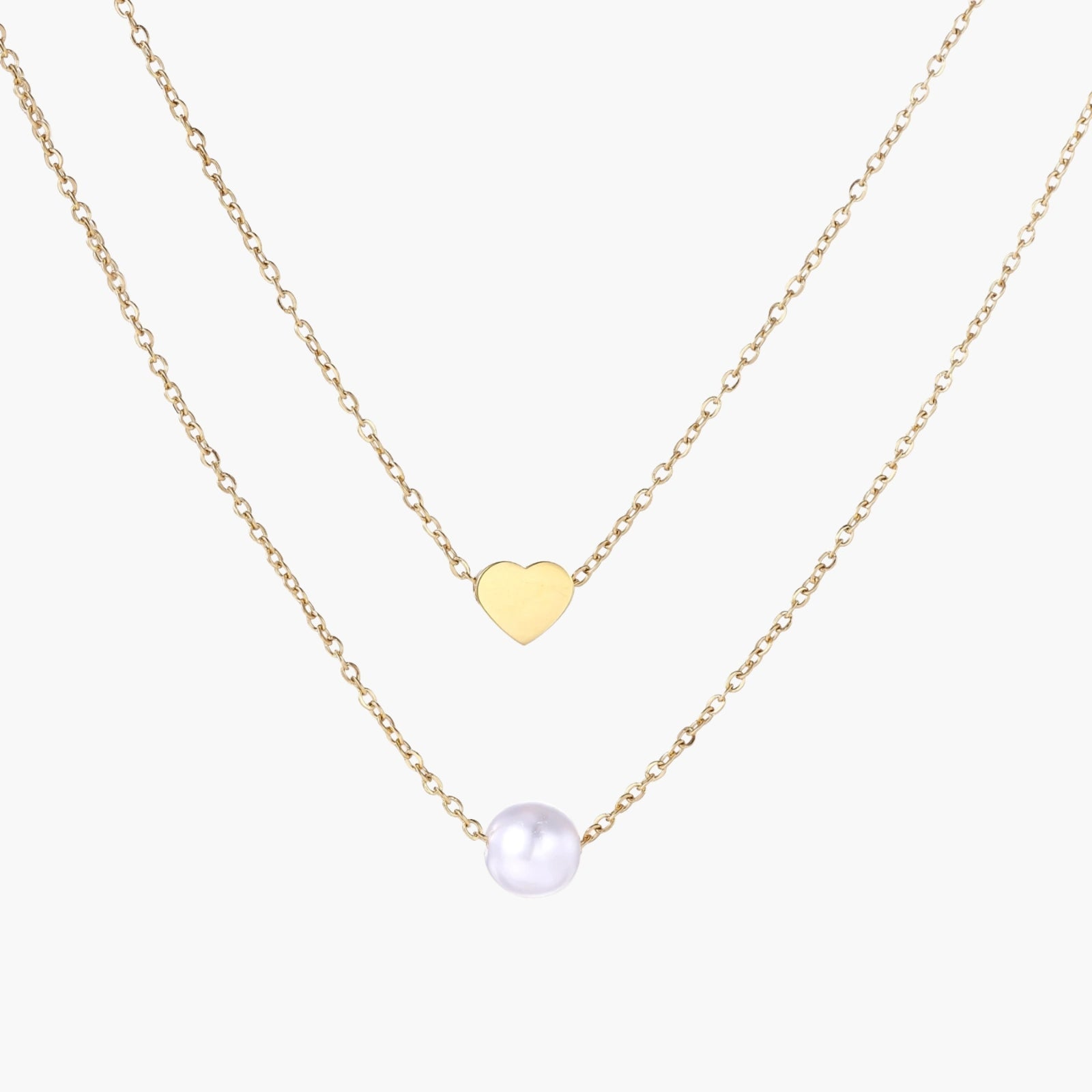 Two delicate gold-plated chains with heart charm and white pearl bead on cream background, minimal layered design.