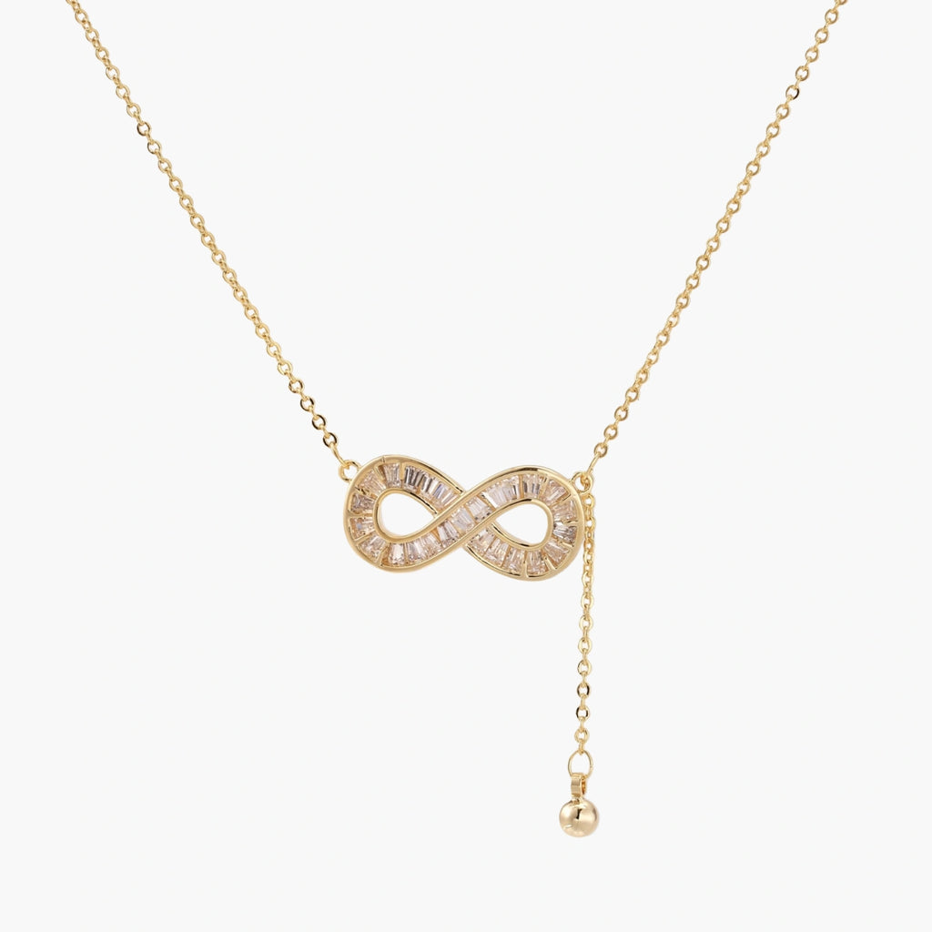 Gold infinity pendant with baguette crystals on adjustable chain against cream background.
