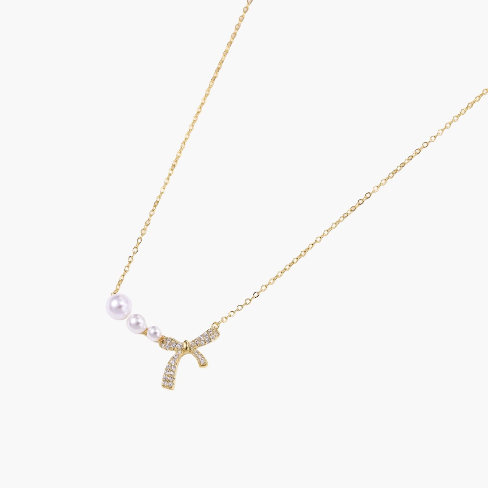 Delicate gold necklace with three white pearls and crystal-embellished bow pendant on cream background