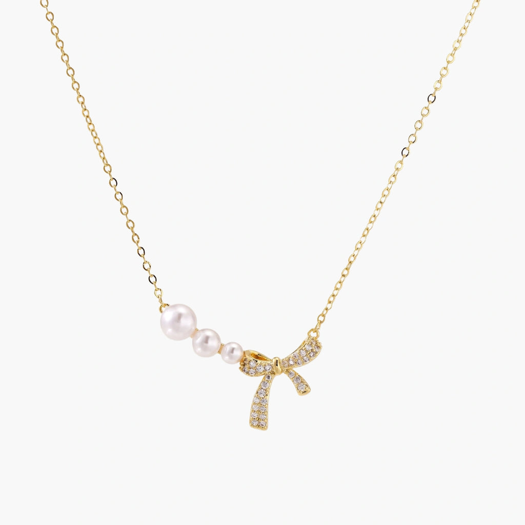 Delicate gold necklace with three white pearls and crystal-embellished bow pendant on cream background